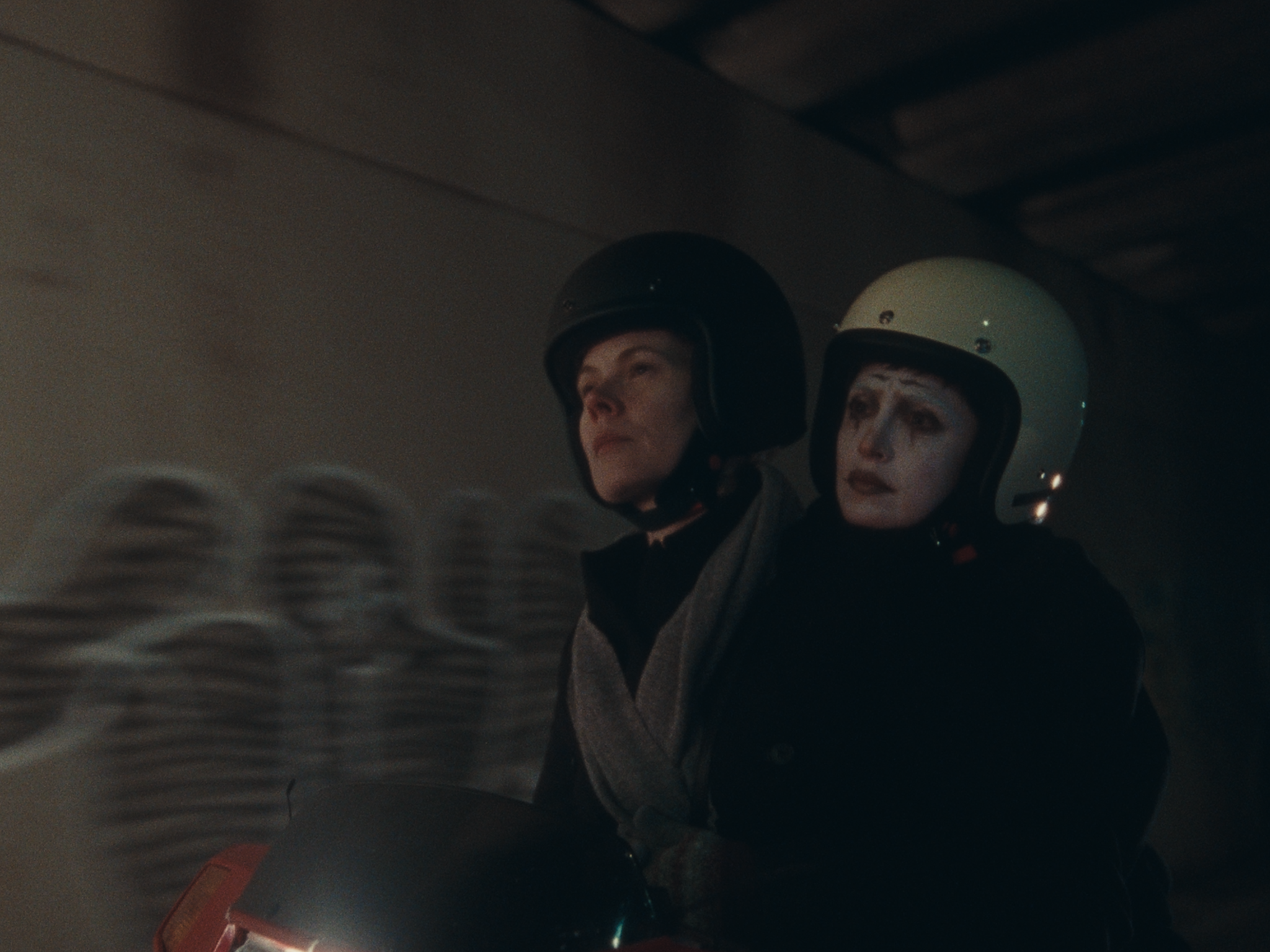 Two characters are seated on a moped, the protagonist looks forlorn and is riding as passenger with their hands clasped around the driver